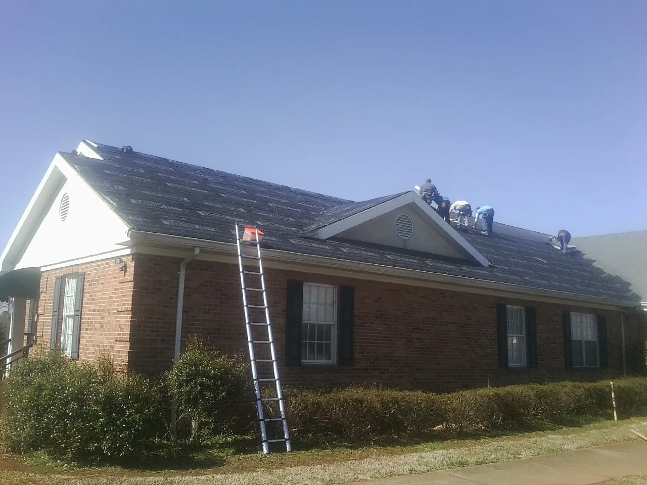 Crew tearing shingles off a brick ranch roof with a ladder set — Emergency Roof Repair work by our crew in Channahon