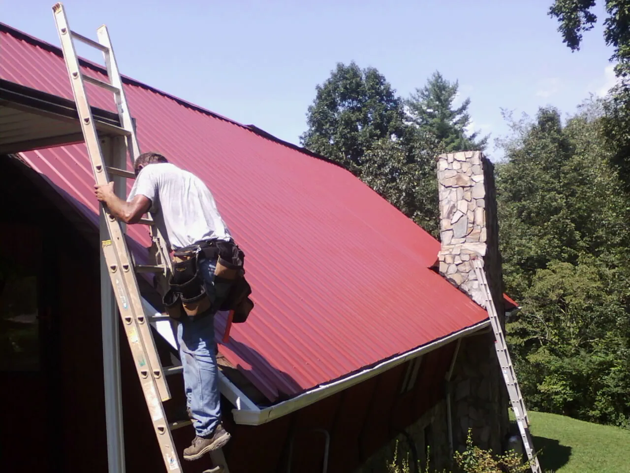 Roofer on a ladder beside a red metal roof and stone chimney — Soffit & Fascia Repair by Roofing Brothers in Channahon